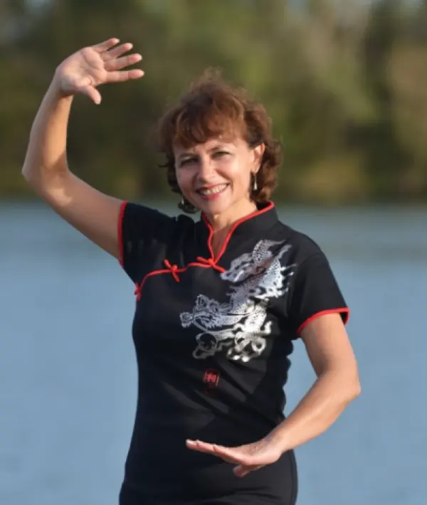 Cours de qi-gong à Mâcon - École des 3 énergies Irène souriante en tenue traditionnelle chinoise noire avec une broderie de dragon argenté, réalisant une posture de qi-gong les bras déployés